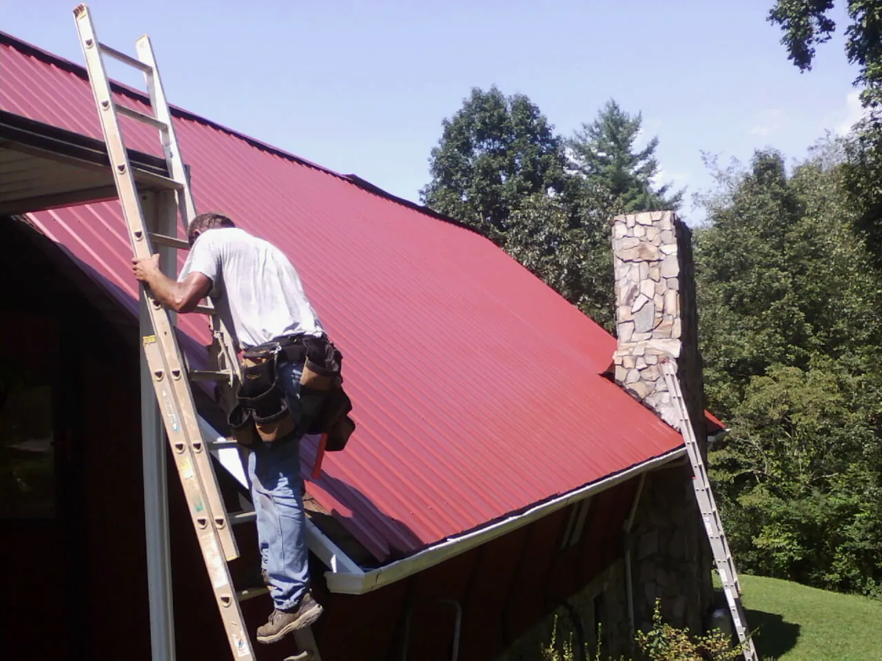 Roofer on a ladder beside a red metal roof and stone chimney — Soffit & Fascia Repair by Roofing Brothers in Bogalusa